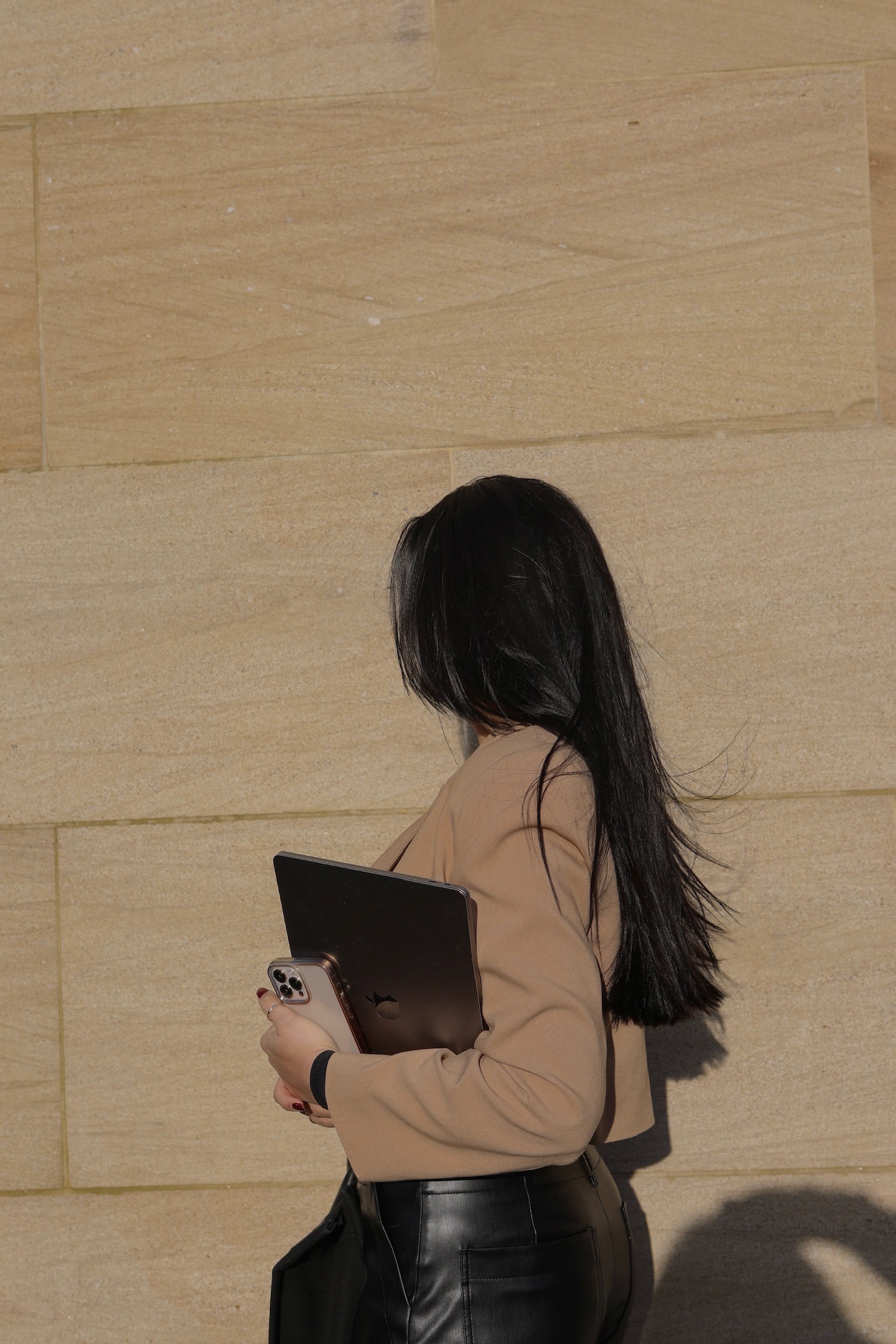 Woman with long dark hair wearing beige sweater and leather pants holding laptop and phone against sandstone wall