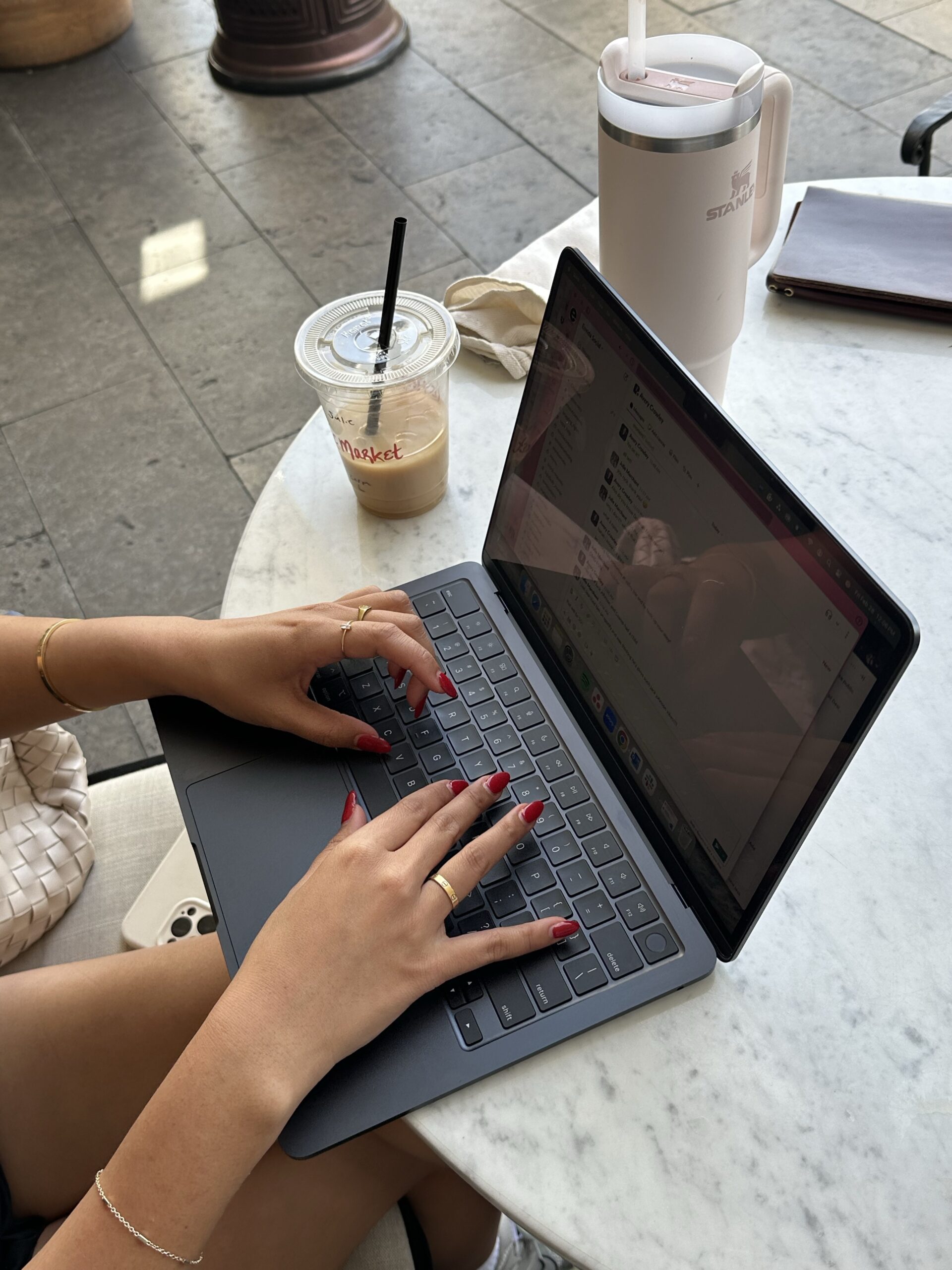 Hands with red manicure typing on laptop at marble cafe table with iced coffee, showing website designer working on Showit platform